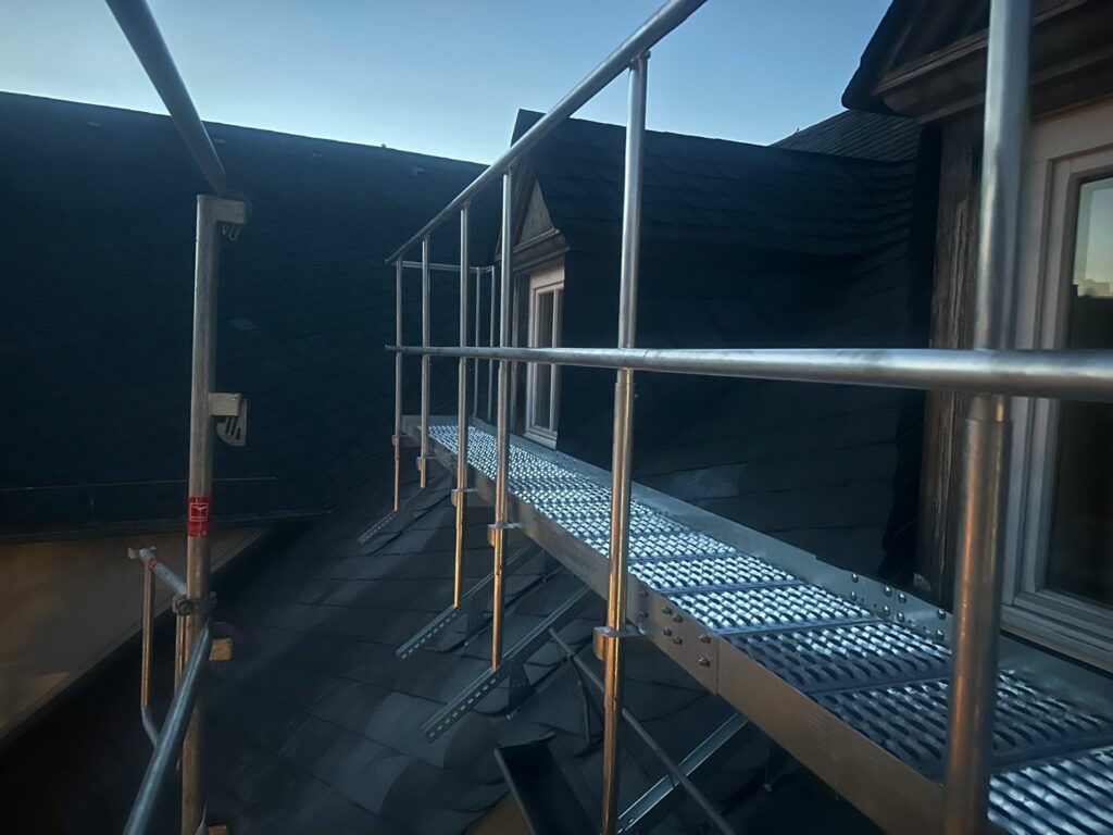 Blick entlang eines langen metallenen Dachlaufstegs mit Geländer vor dunklem Schieferdach.